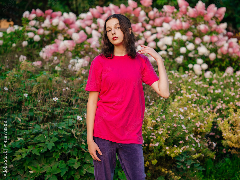 A young woman in a pink shirt stands in a lush green garden. Concept of tranquility and beauty, as the woman poses in front of the vibrant flowers and greenery. Model with slim sport body type.