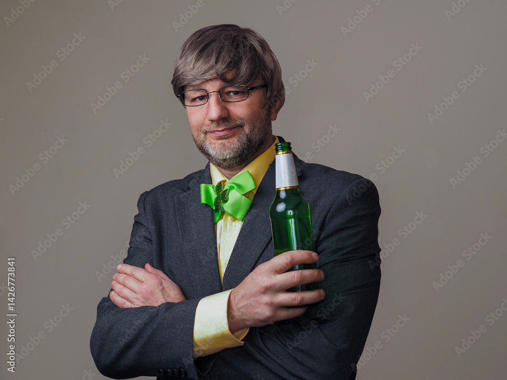 Man in light color shirt and dark jacket and green ribbon with shamrock on a light color background wall. Irish sport fan and support. Male, in his 50s with grey beard and hair. Holding beer bottle.