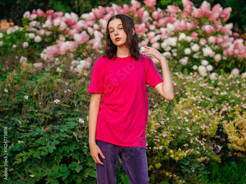 Obraz na płótnie A young woman in a pink shirt stands in a lush green garden. Concept of tranquility and beauty, as the woman poses in front of the vibrant flowers and greenery. Model with slim sport body type. w salonie