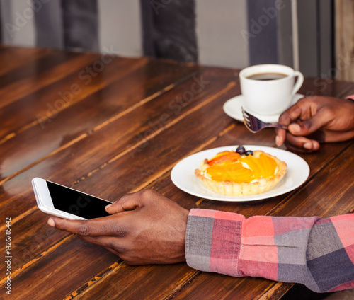 African American man holding smartphone and spoon over fruit tart and coffee cup on café table