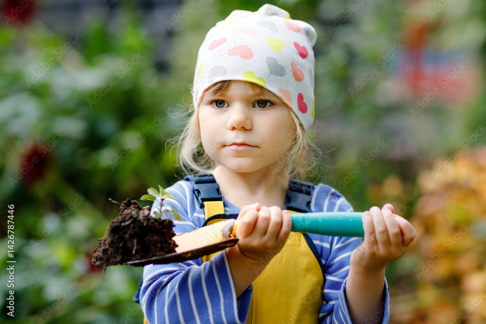 Adorable little toddler girl holding garden shovel with green plants seedling in hands. Cute child learn gardening, planting and cultivating vegetables in domestic garden. Ecology, organic food.