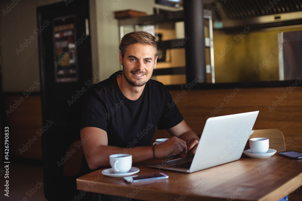 Man in late twenties sitting at modern café table working on silver laptop with coffee cups Man in late twenties sitting at modern café table working on silver laptop with coffee cups