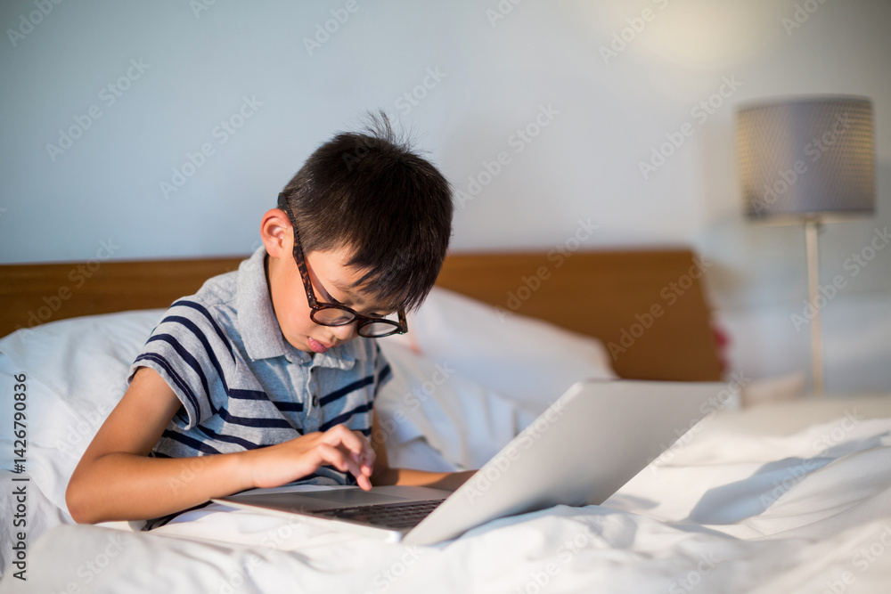 Asian boy child sitting on bed using silver laptop in bedroom with eyeglasses and lamp Asian boy child sitting on bed using silver laptop in bedroom with eyeglasses and lamp