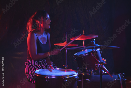 Young adult Asian female drummer performing on stage with drum kit under red and blue lights