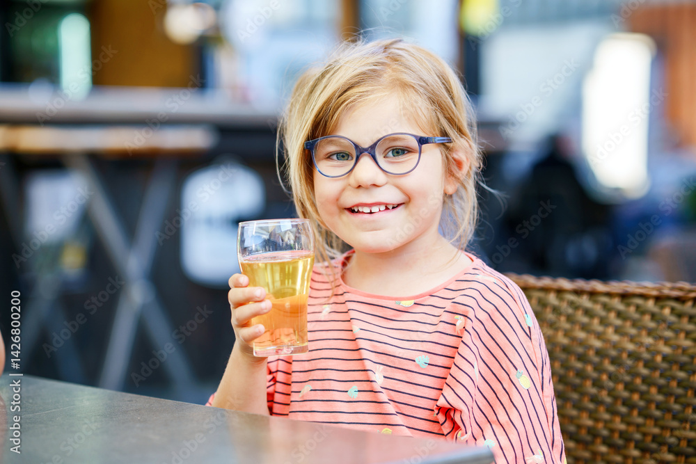 Adorable little girl having breakfast and drinking juice at resort restaurant. Happy preschool child with glasses holding glass with apple juice