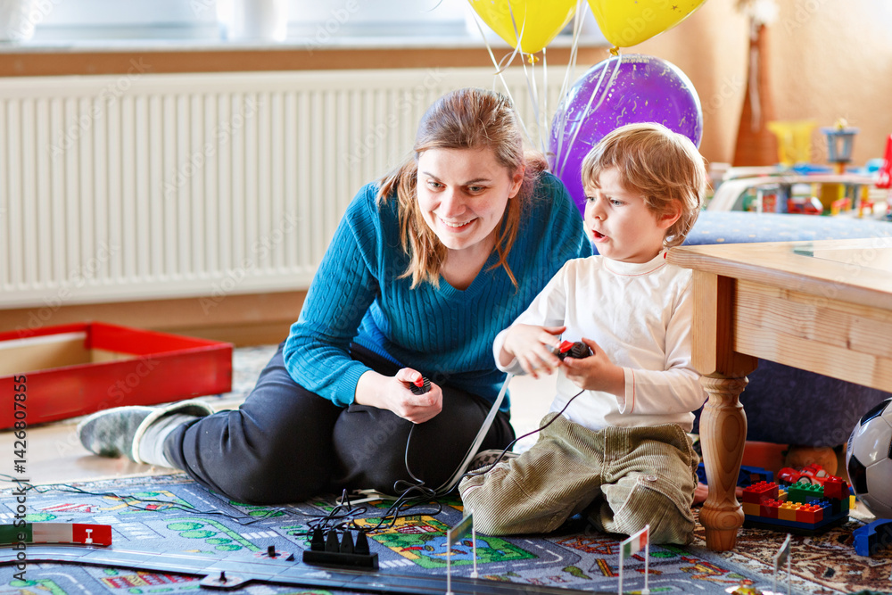 Mother and son playing with racing cars on racetrack, indoors, with cars. Happy little preschool boy and young woman having fun with indoor race game