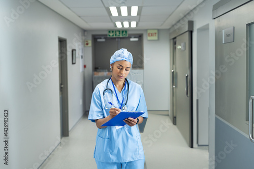 Obraz na płótnie African American woman standing in hospital corridor, writing on blue clipboard with pen w salonie