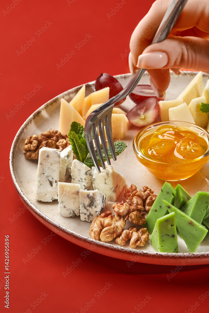 Woman using fork taking gorgonzola cheese from plate with variety of cheeses, grapes, walnuts and honey on red background