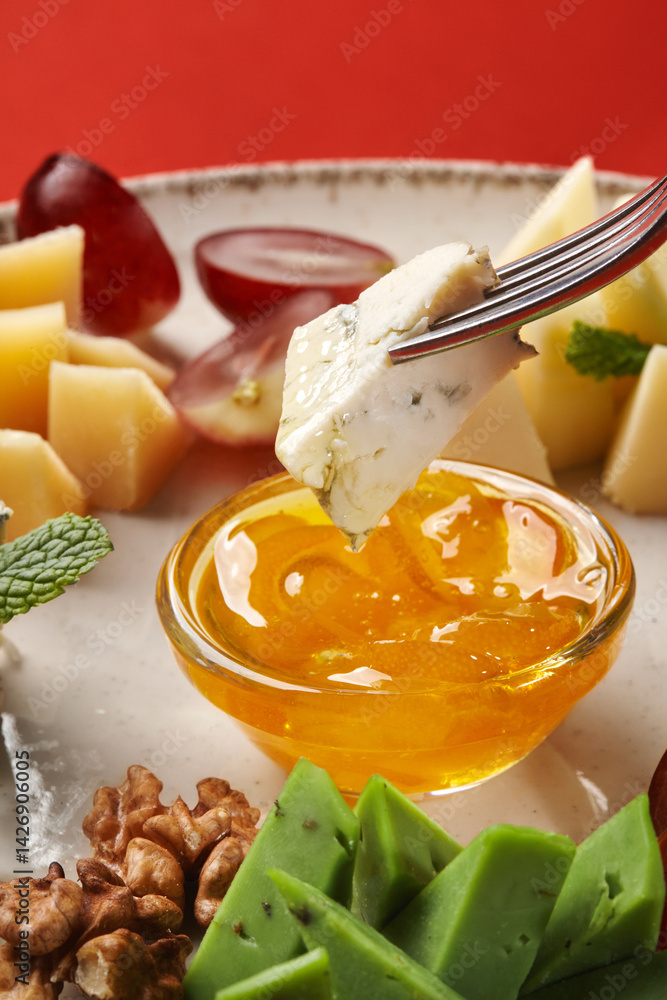Fork dipping blue cheese into honey with variety of cheeses, nuts, and grapes on platter