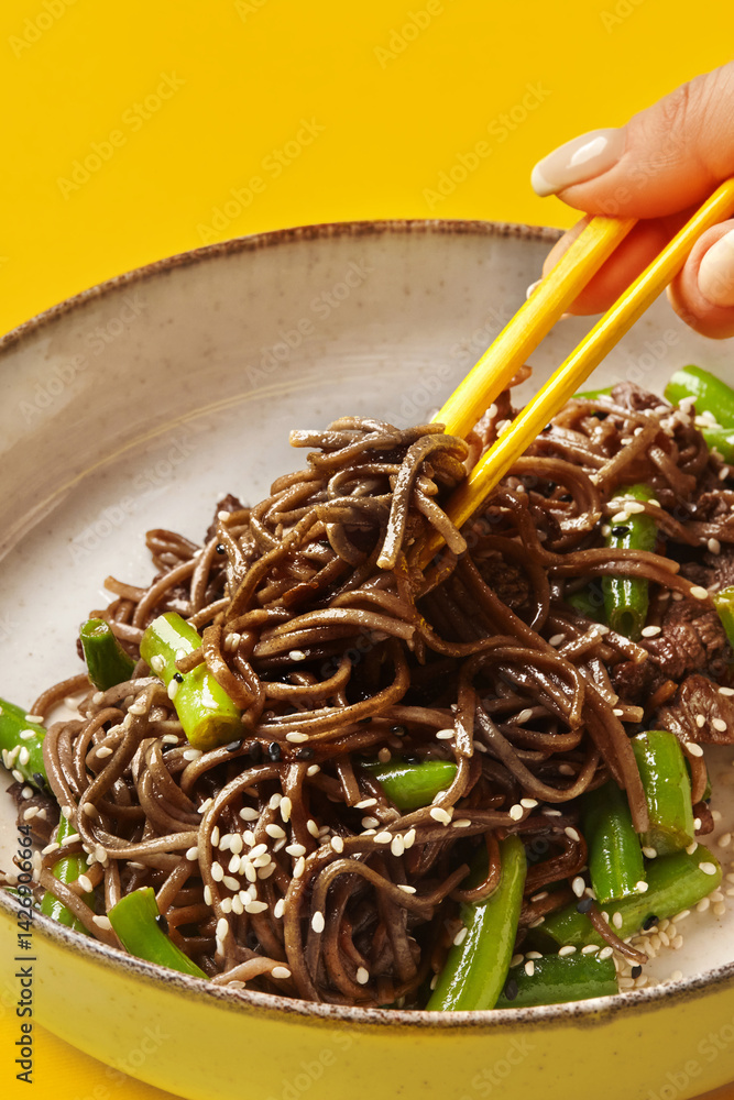 Woman using chopsticks eating soba noodles with green beans and sesame seeds on yellow background
