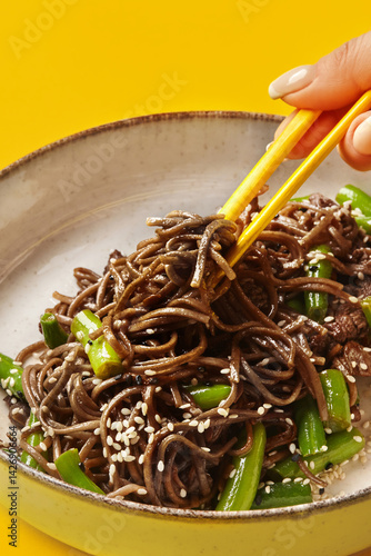 Obraz na płótnie Woman using chopsticks eating soba noodles with green beans and sesame seeds on yellow background w salonie