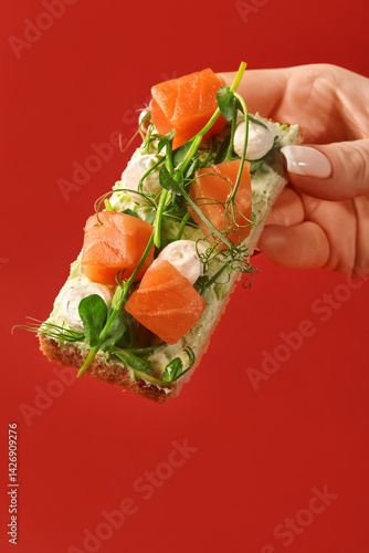 Obraz na płótnie Woman hand holding tasty bruschetta with salmon, cream cheese and microgreens on red background w salonie