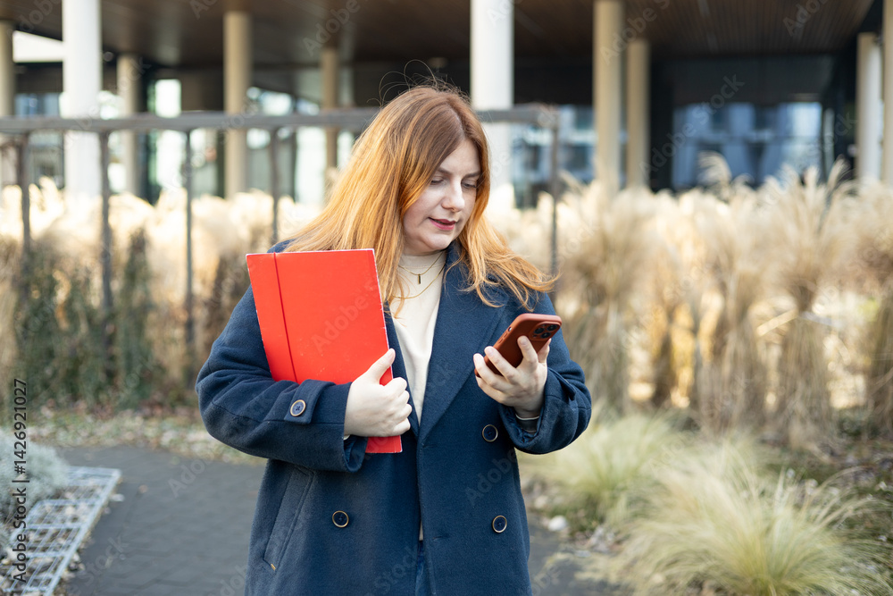 Young adult smiling professional business woman holding smartphone and folder with files while walking in urban city street.