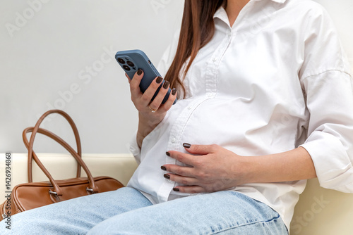 Obraz na płótnie Unrecognizable pregnant woman using a smartphone and gently touching her belly while relaxing on couch in hospital. Happy young expectant mother. Family planning and IVF centers concept. High quality w salonie
