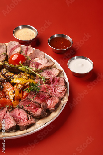Obraz na płótnie Sliced beef and pork tenderloin with grilled vegetables and sauces on red background w salonie