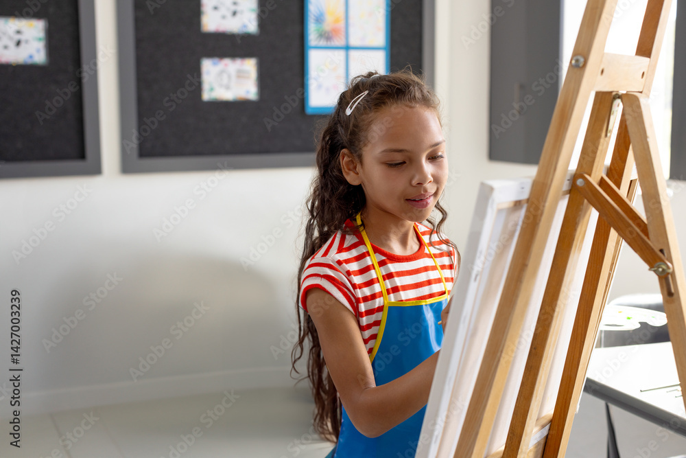Female child painting on canvas on easel in art classroom, with paintbrush and pinned drawings