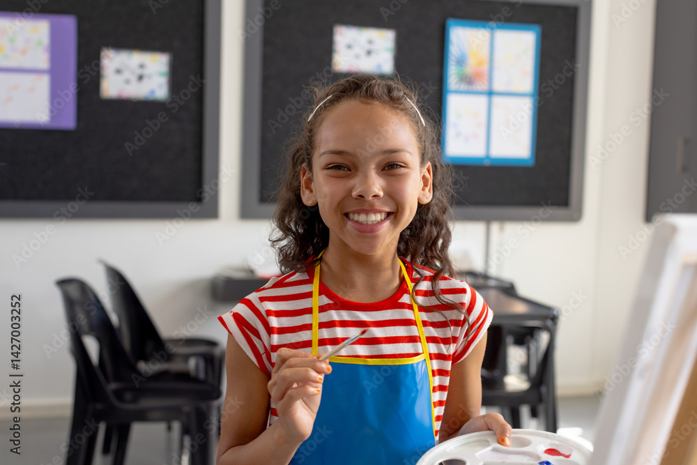 Multiracial school-aged girl painting in school art classroom, holding palette and paintbrush