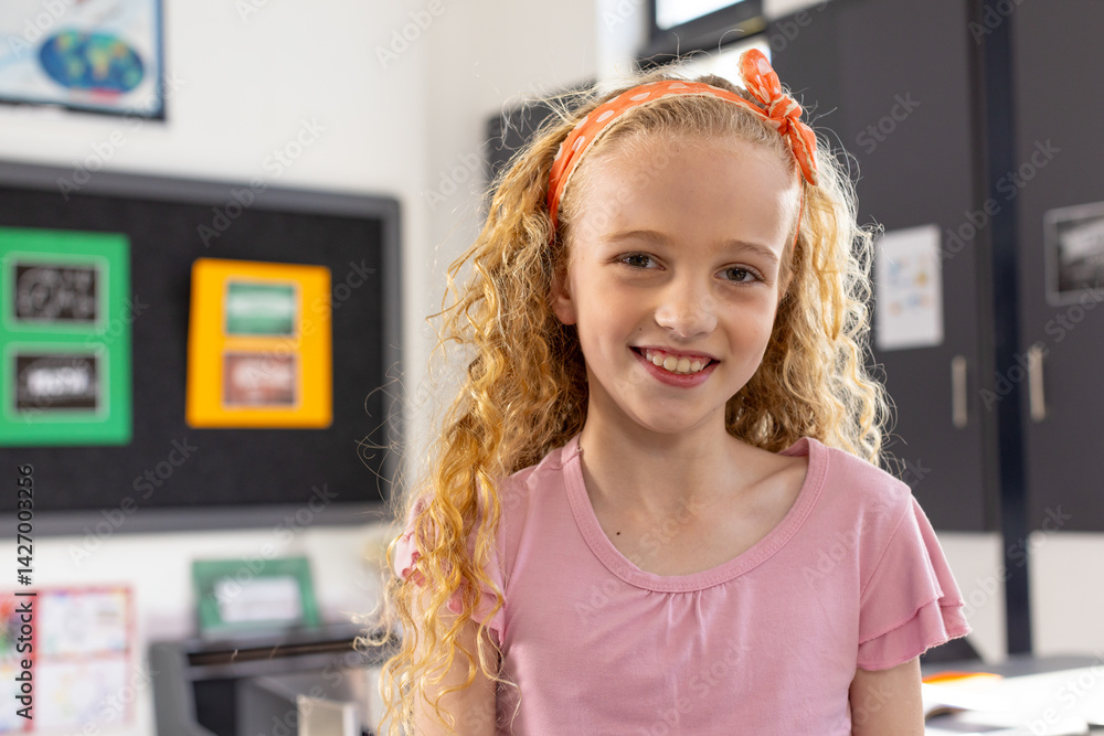 girl child standing and smiling in bright classroom, with bulletin board, world map and desk books