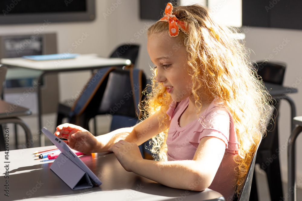 Female child student using tablet on folding stand at school desk in bright classroom, copy space