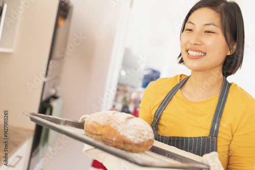 Obraz na płótnie Asian woman holding freshly baked loaf on metal baking tray in home kitchen, copy space w salonie