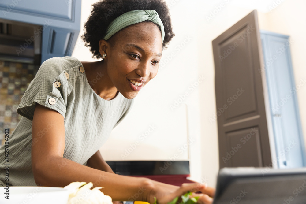 Young African American woman leaning on kitchen counter chopping herbs and using tablet, copy space