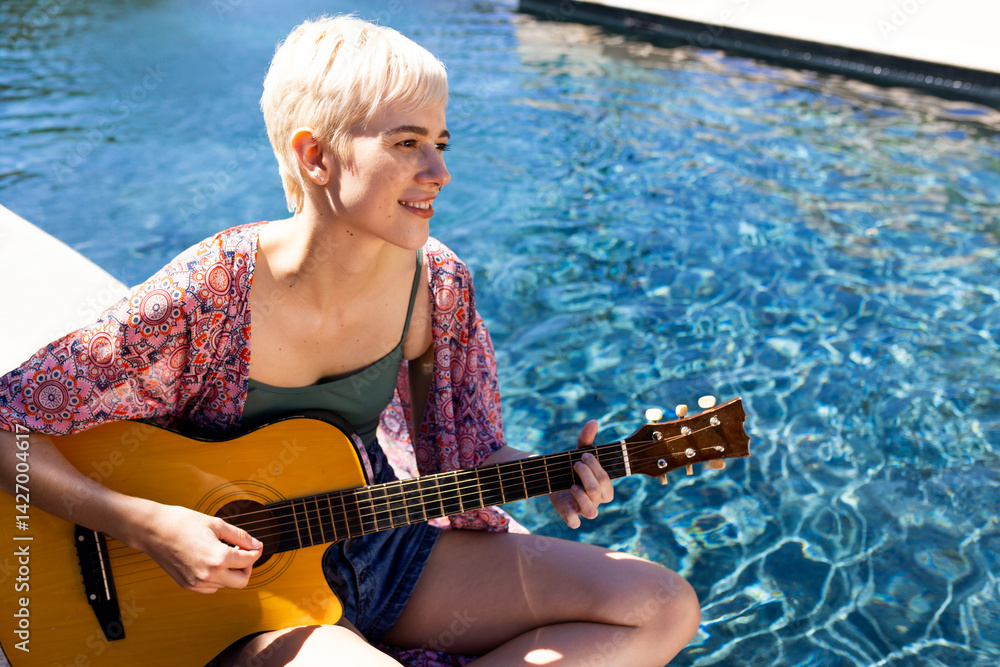 Non-binary musician sitting poolside in sun, playing acoustic guitar near clear water, copy space