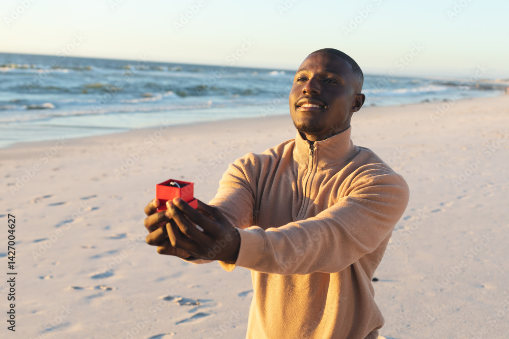 African American man kneeling on sandy shore, presenting red box holding diamond ring, copy space