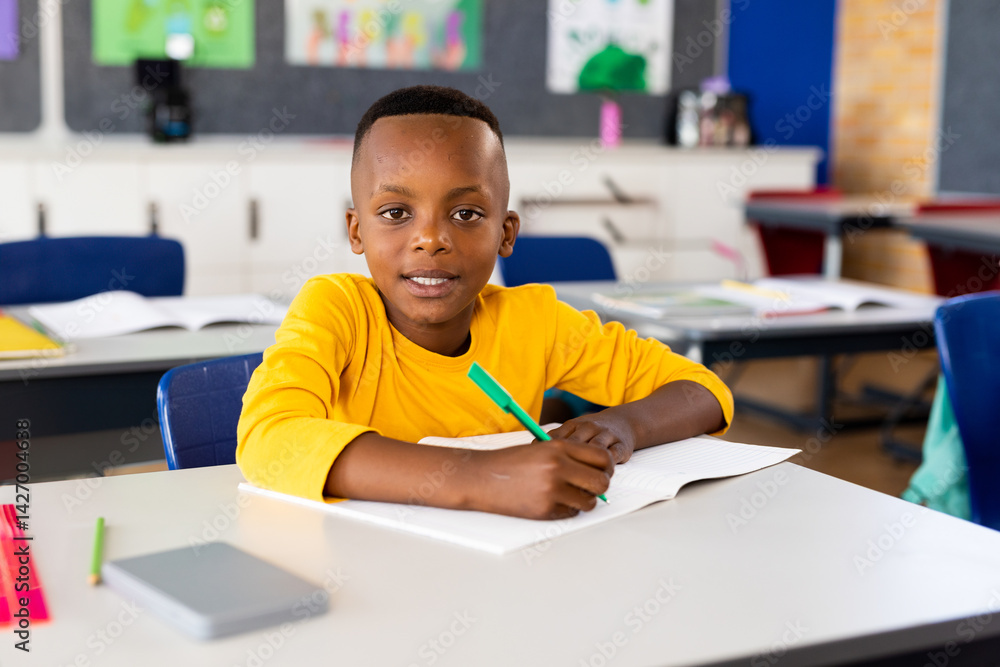 African American child writing in open notebook at school desk, using green pen and smiling