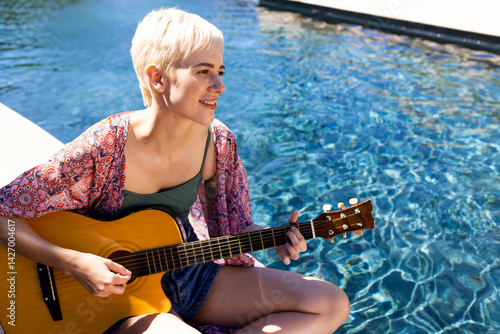 Obraz na płótnie Non-binary musician sitting poolside in sun, playing acoustic guitar near clear water, copy space w salonie
