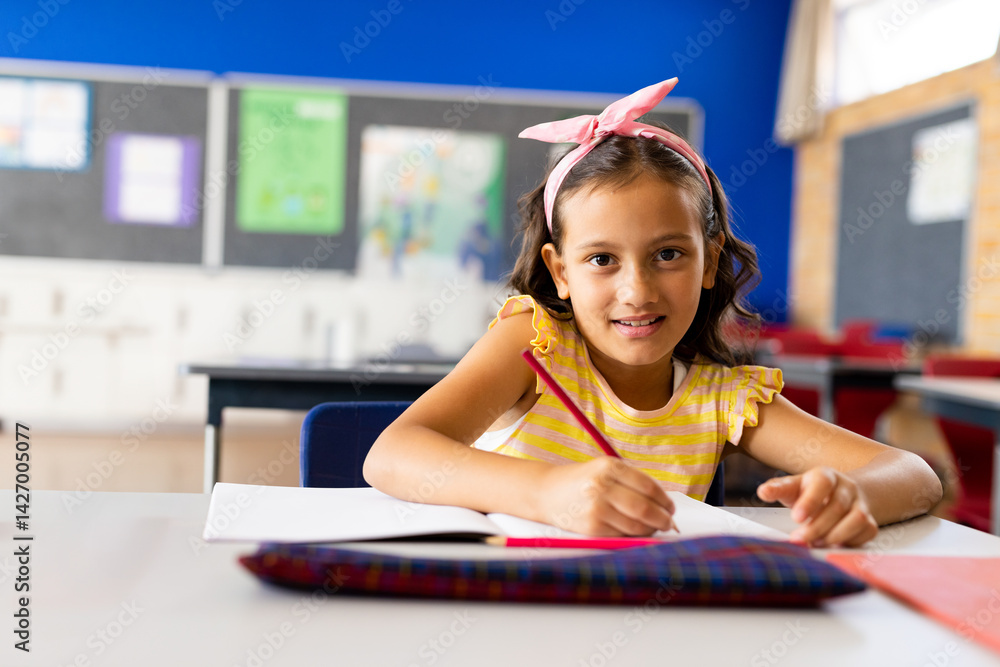 Child girl student writing in elementary classroom with blue wall, open notebook and red pencil