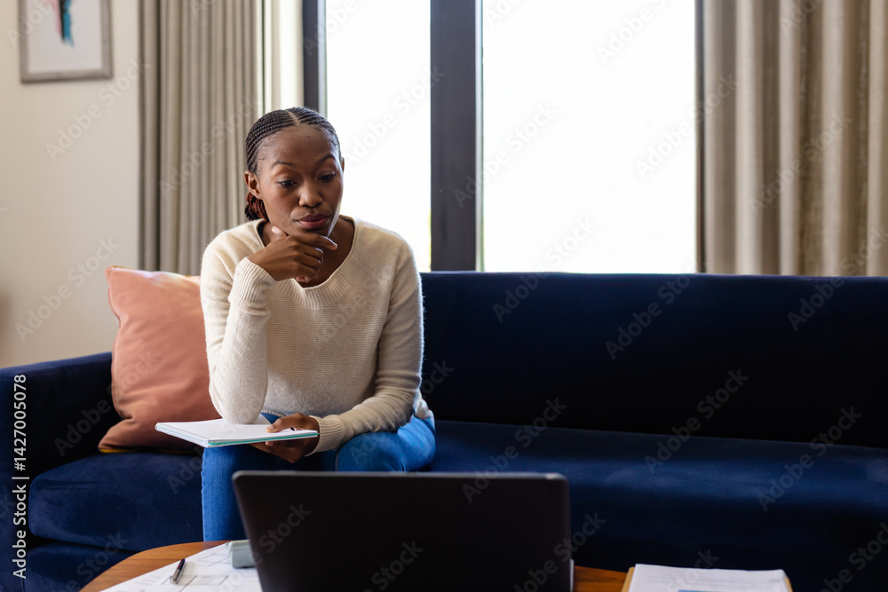 African American woman studying on sofa in living room, with laptop, notebook, pen, copy space