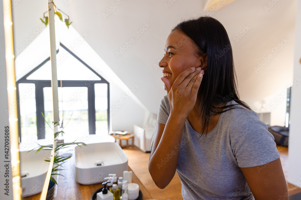 African American woman applying cream in attic bathroom by mirror, with skincare jars on counter