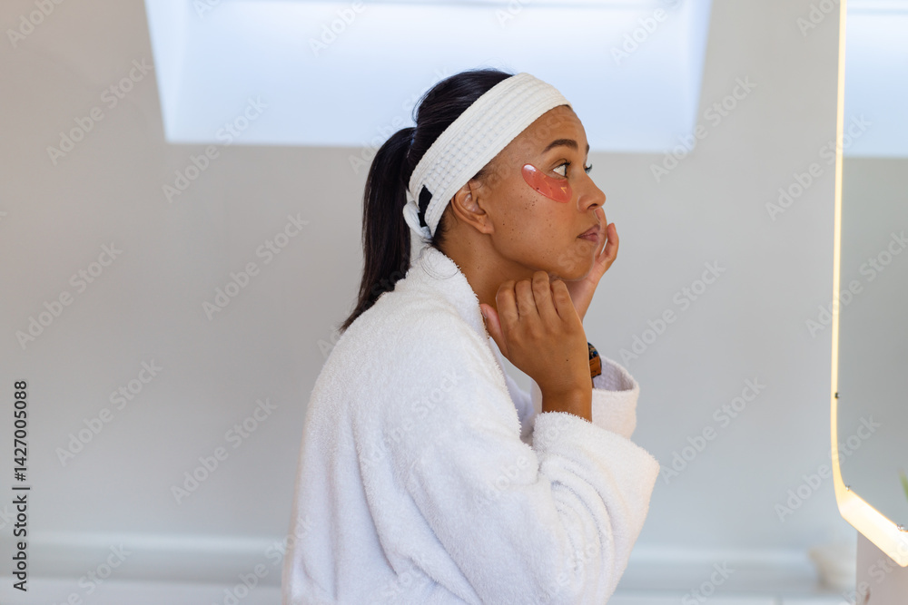 African American woman applying pink under-eye patch in bathroom, facing backlit mirror, copy space