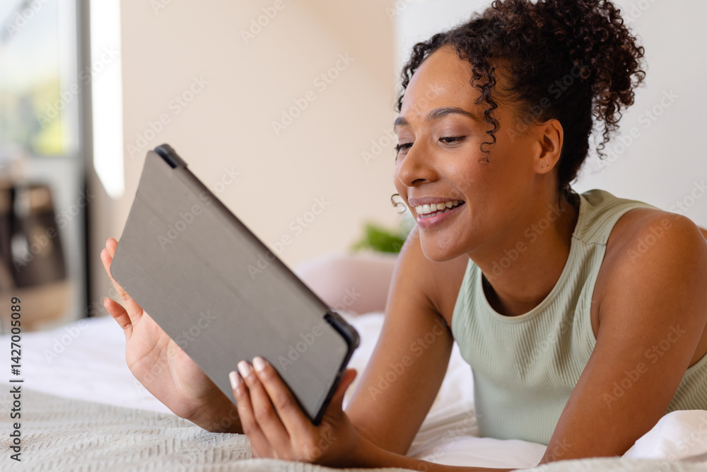 Diverse young woman lying on stomach on bed in bright bedroom, holding tablet, smiling, copy space