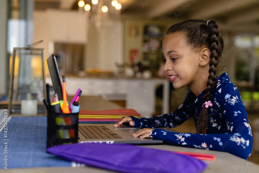 Child girl sitting at table using laptop in home kitchen, with pen holder, notebooks, copy space