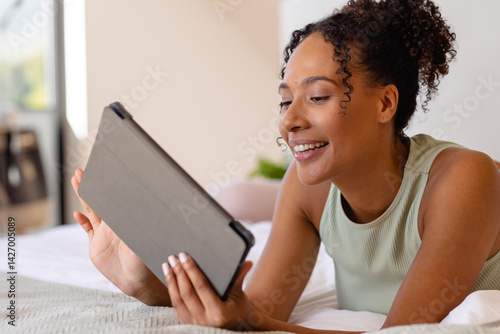 Obraz na płótnie Diverse young woman lying on stomach on bed in bright bedroom, holding tablet, smiling, copy space w salonie