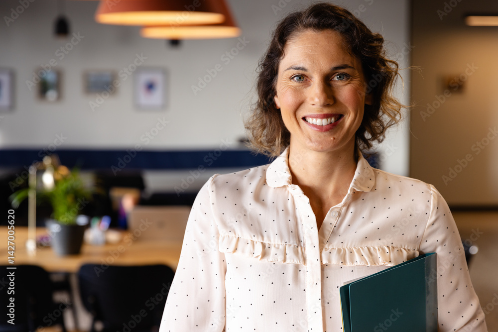 Woman standing and smiling while holding green folder in coworking office with wooden desk, laptop