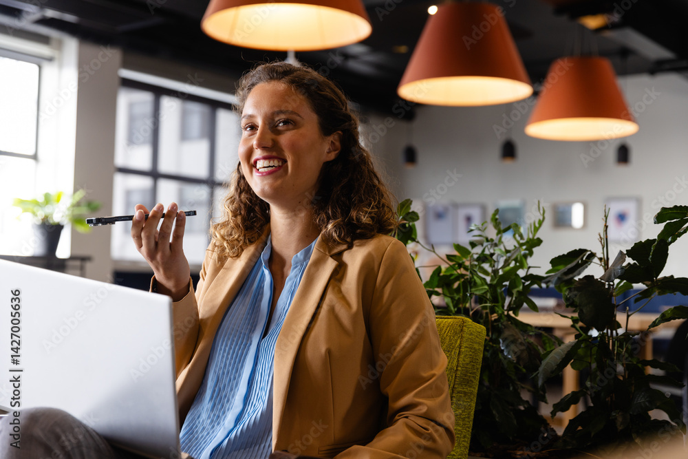 Young adult woman speaking on smartphone at desk in open-plan office lounge, with laptop and plants