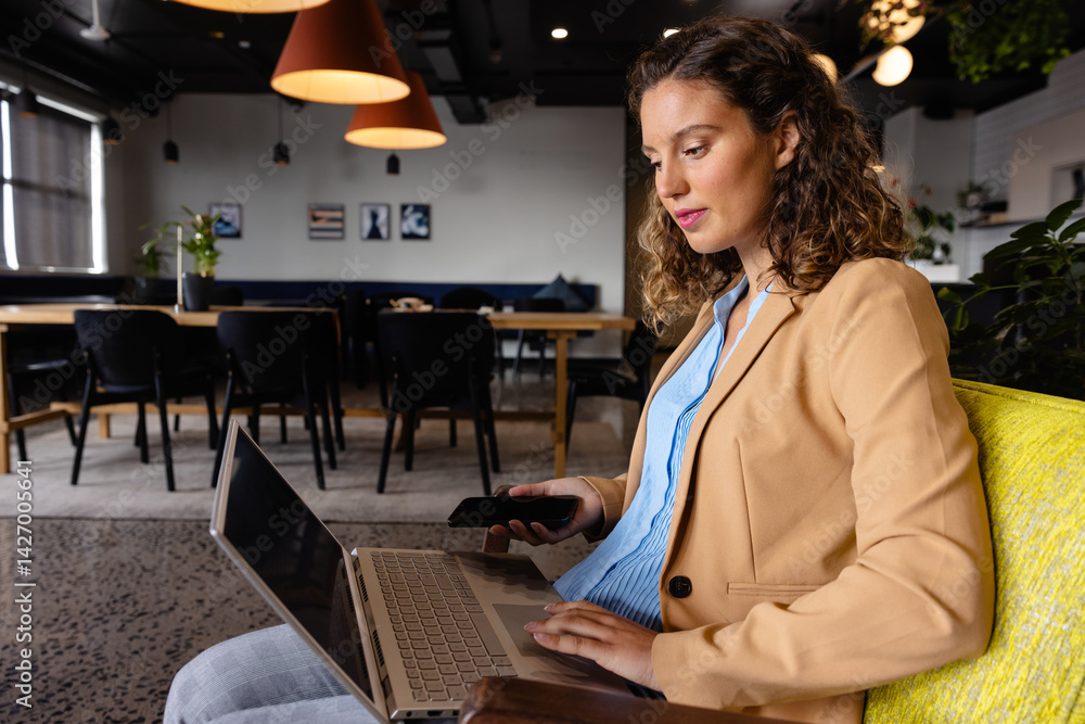 Young woman using laptop and smartphone in chartreuse armchair in coworking space, copy space
