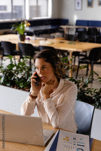 Woman talking on smartphone while using silver laptop at open-plan office desk with charts
