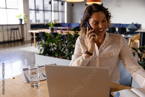 Obraz na płótnie Woman smiling and talking on smartphone while using laptop at open plan office desk, copy space w salonie