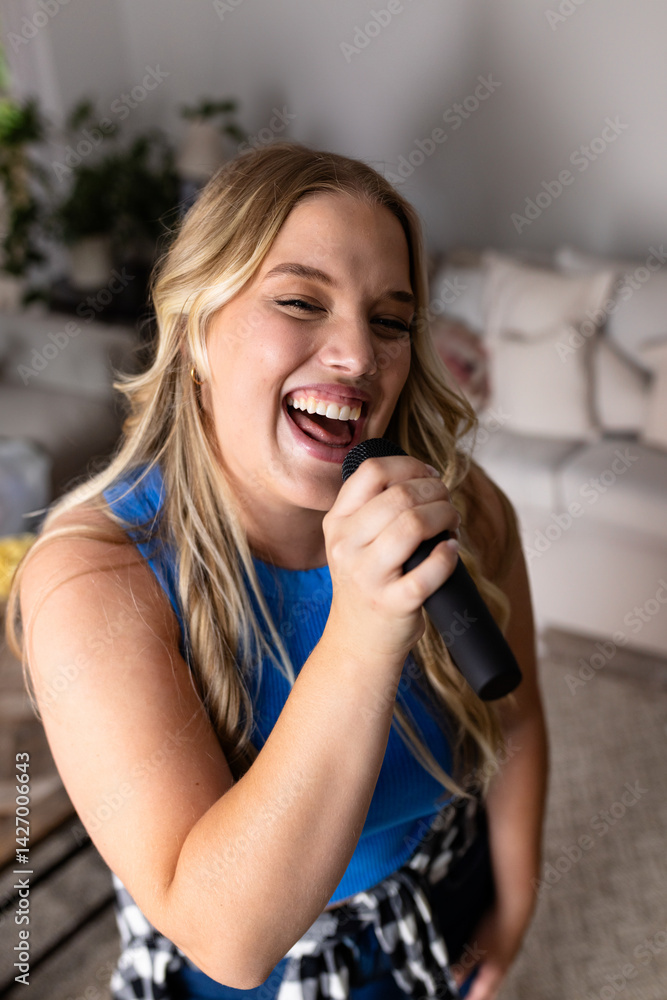 Young adult woman singing while standing in living room, holding handheld microphone and smiling