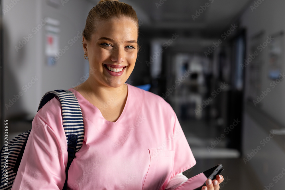 Young adult nurse wearing pink scrubs and striped backpack in hospital corridor, holding smartphone