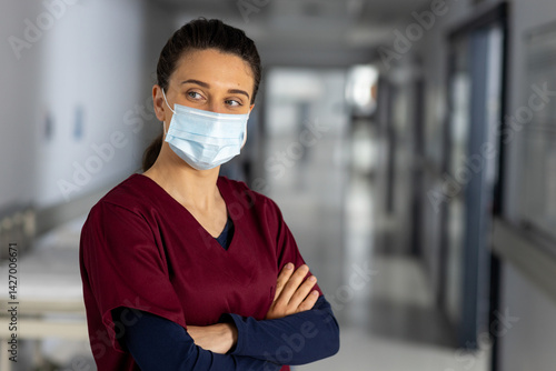 Young female nurse standing in hospital hallway with burgundy scrubs and blue mask, copy space