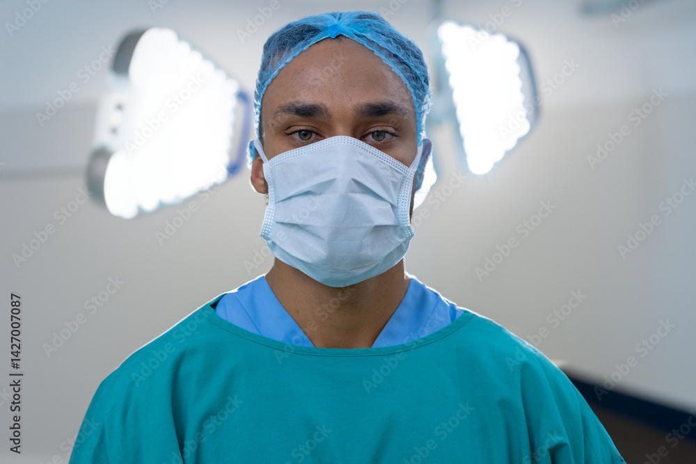 Male professional standing in operating room under LED lights, wearing surgical gown, cap and mask