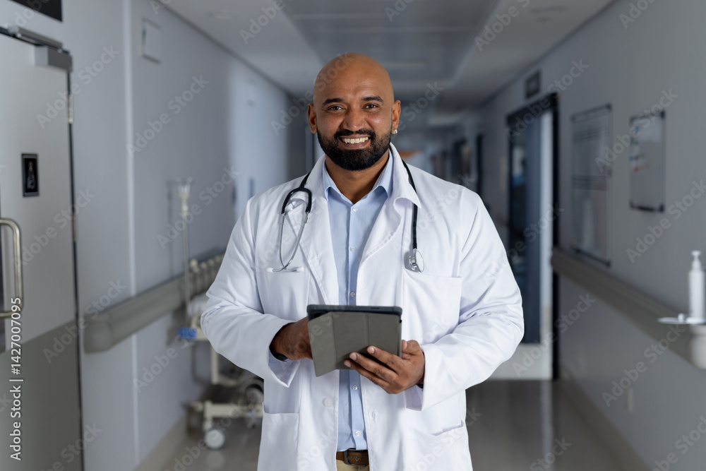 Male doctor holding tablet and smiling in hospital corridor, with stethoscope and IV stand