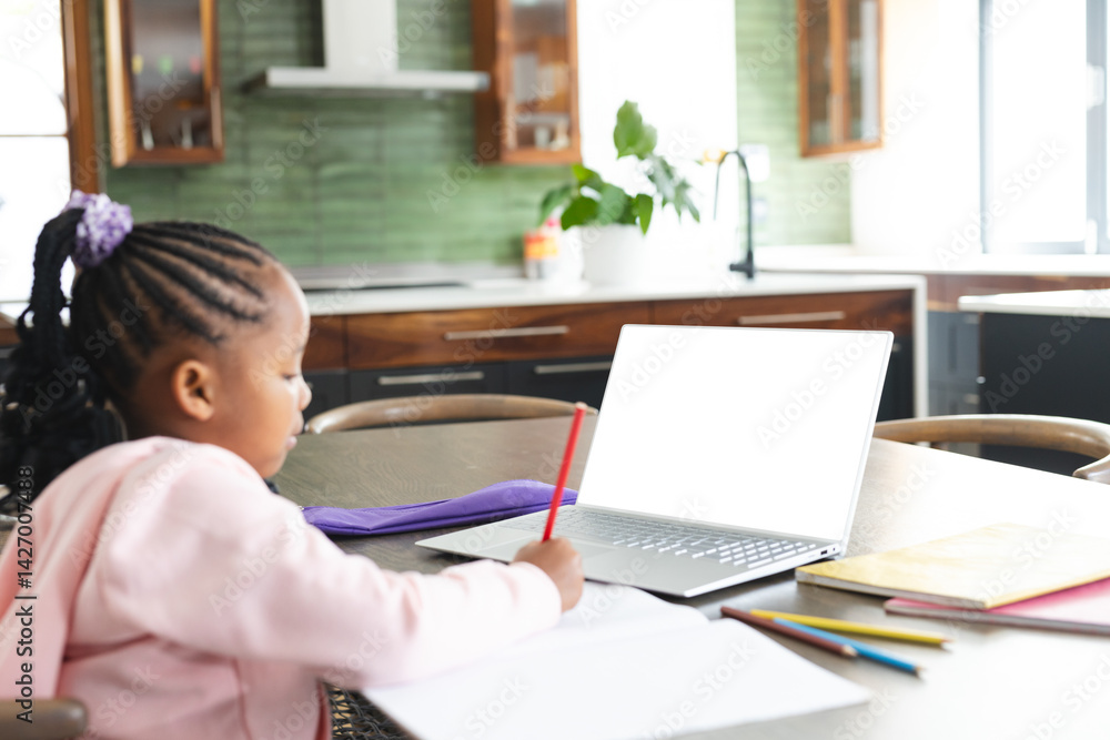 African American girl using silver laptop and writing with red pencil at kitchen table, copy space