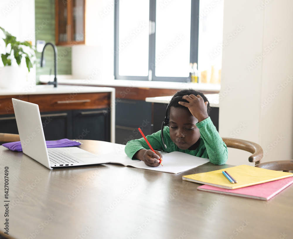 African American boy writing in notebook at table in modern kitchen, with laptop and headphones