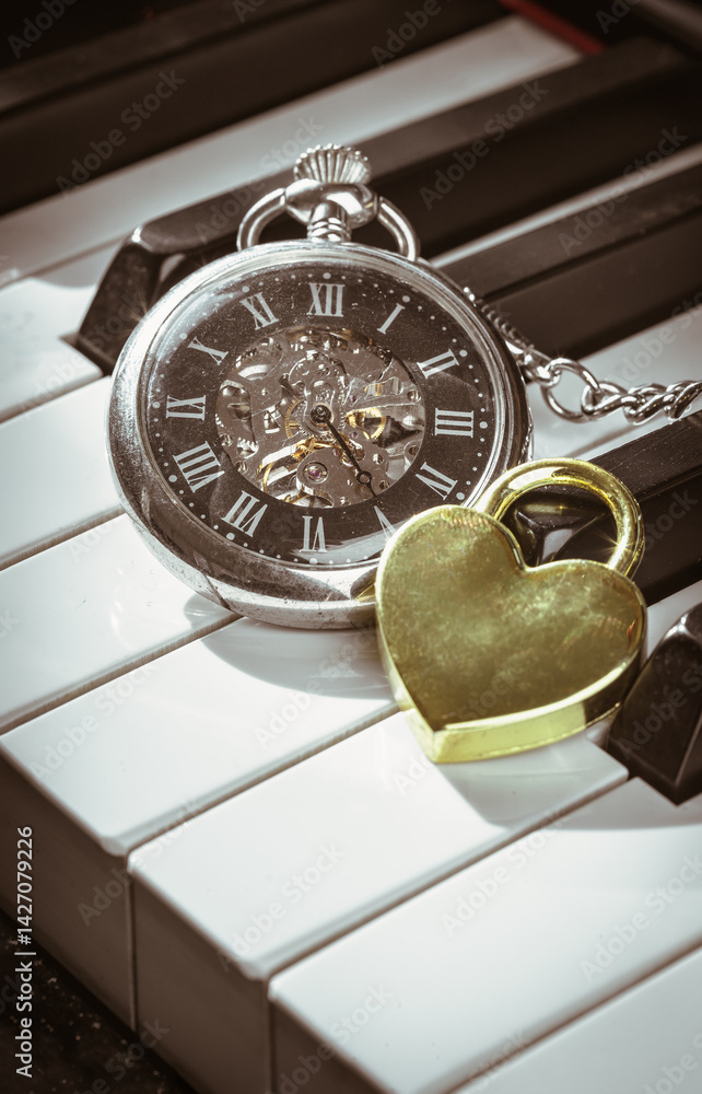 A retro pocket watch or Antique pocket watch with Golden heart shaped love padlock on Piano keys background in Vintage tone. The concept of the time of love, use it as your Wallpaper, Poster and Space
