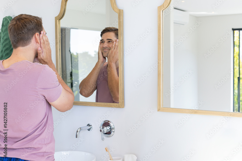 Man touching face at sink mirror in bright modern bathroom, with toothbrush and towel, copy space